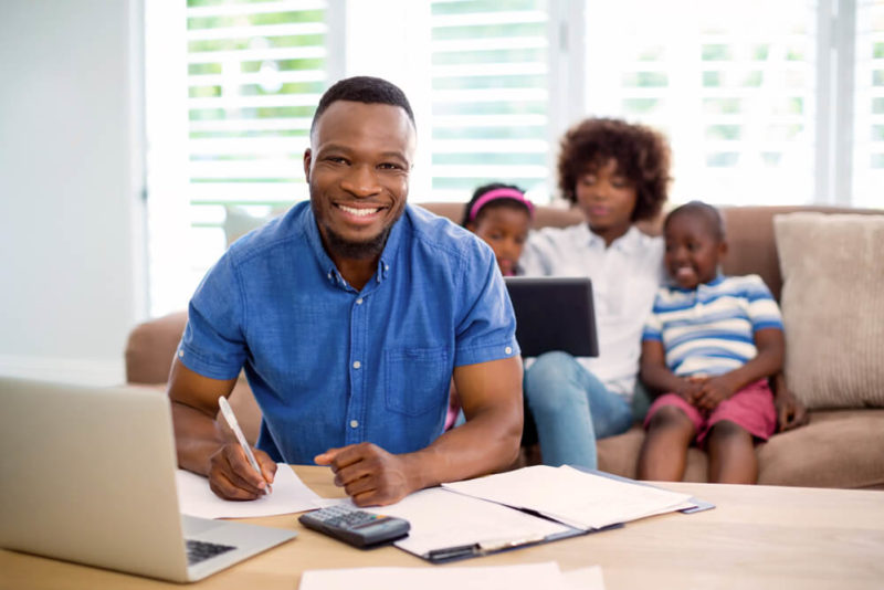 Smiling man calculating in front of a laptop with family wife and kids in teh background sitting on the couch.