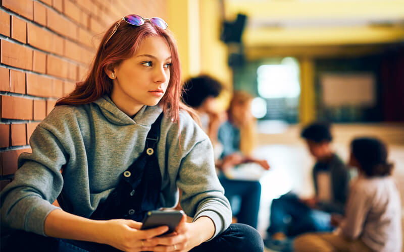Teenage girl at school sitting on the floor in the hallway with her phone looking away.