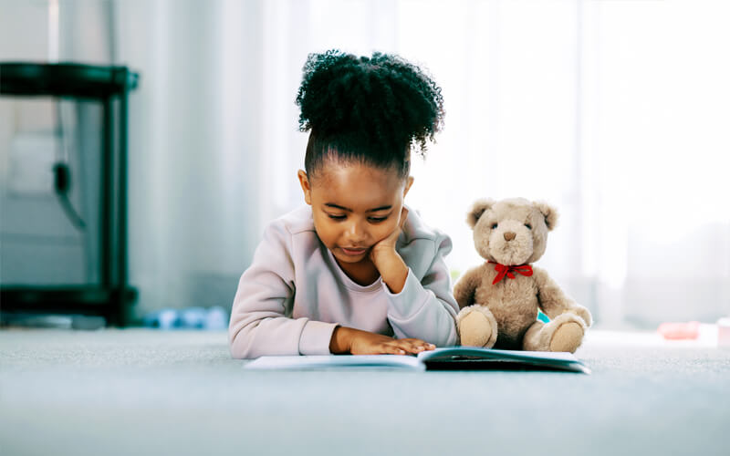 Young girl reading her book on floor with her teddy bear.