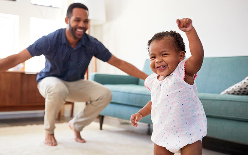 Baby daughter dancing with father In lounge at home