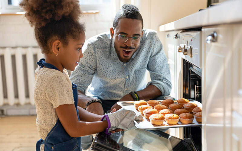 Excited african american dad watching daughter taking muffins out of oven