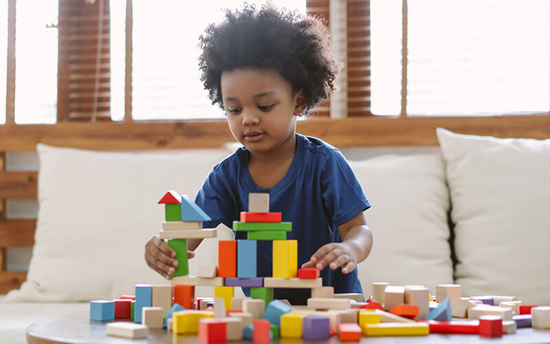 Little African-american boy building a small house with colorful wooden blocks in the living room at home