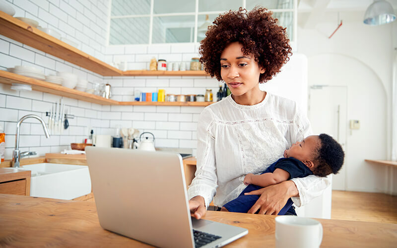 Multi tasking mother holds sleeping baby son and works on laptop computer in the kitchen